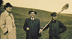 Three individuals stand in an open field; one holds a shovel.
