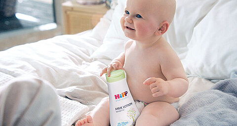 A baby sits on a bed, holding a bottle of Hipp Milk Lotion.