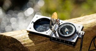 A compass and a lens cap are placed on a wooden log with a blurred background of water droplets.